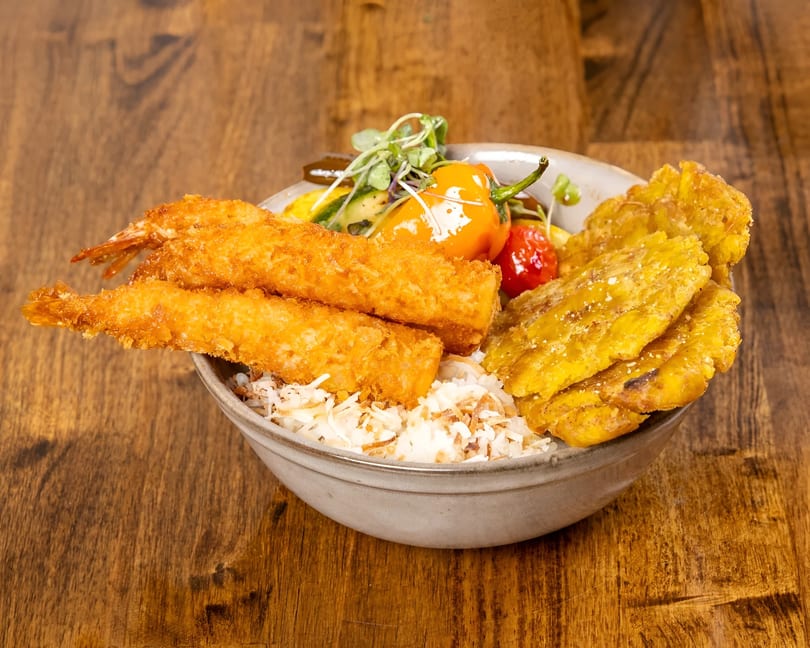 A colorful bowl of fried shrimp, plantains, rice, and vegetables on a wooden table.