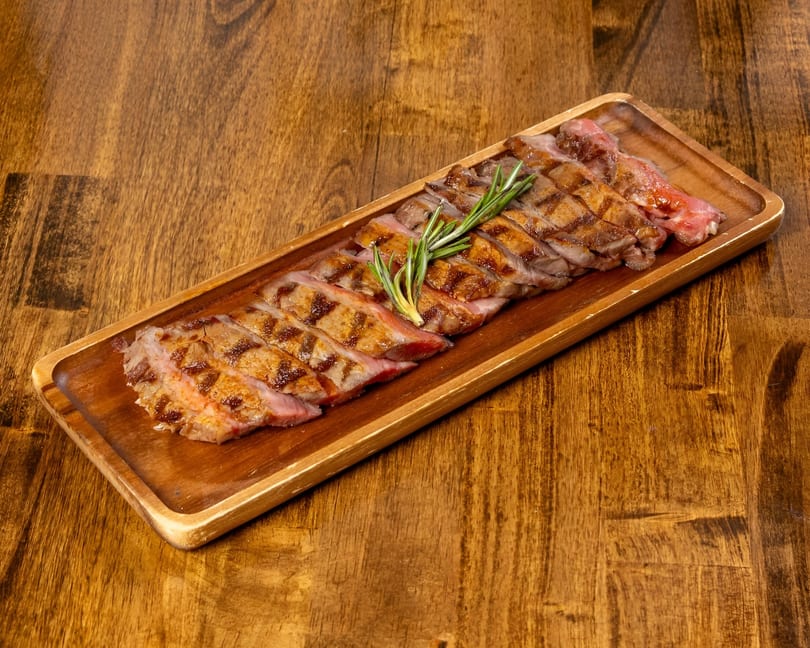 Grilled steak slices with grill marks and rosemary sprigs on a long wooden serving board.